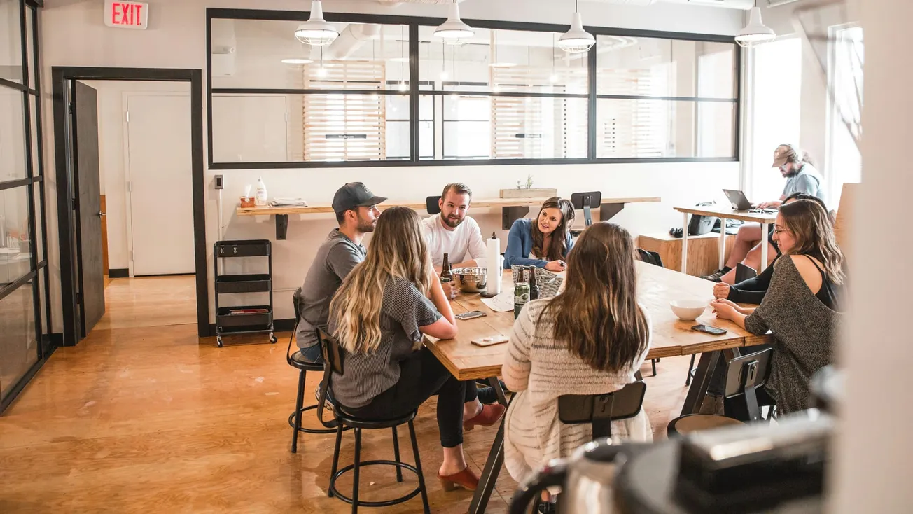 A group of seven people sit around a wooden table in a modern office, engaged in a meeting. Large windows and hanging lights create a bright, collaborative atmosphere.