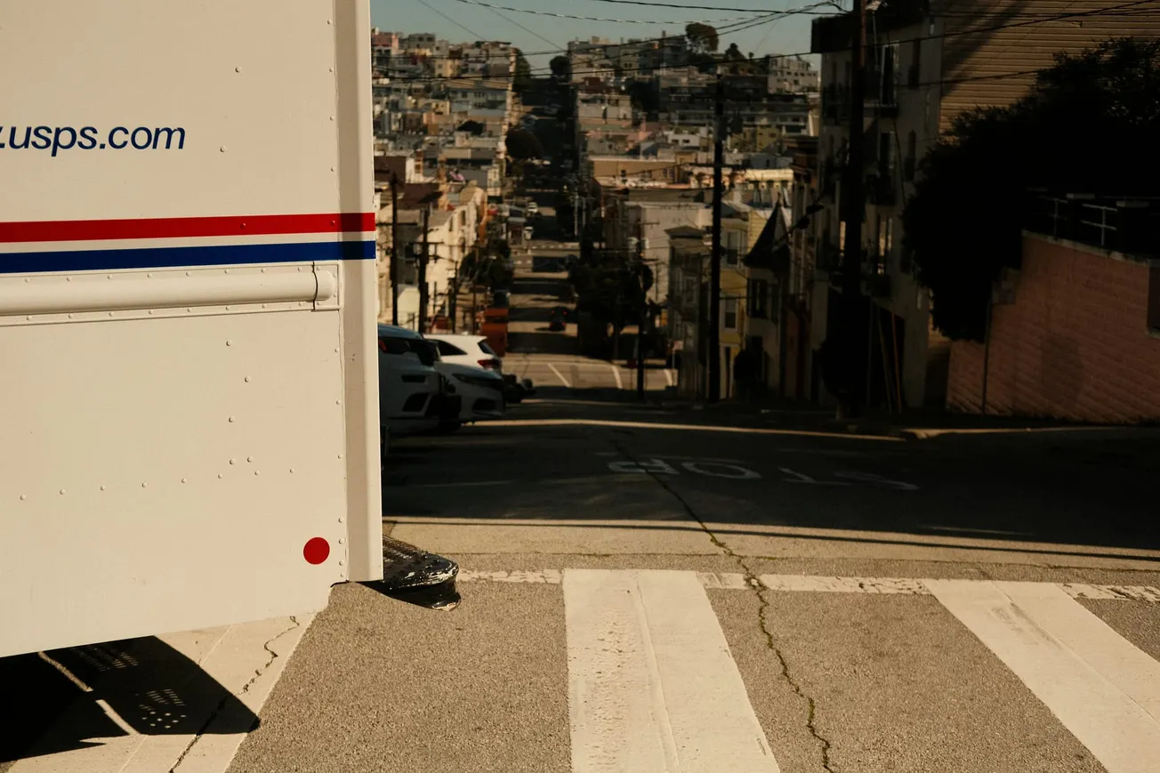 USPS truck parked near a crosswalk on a steep street, with a distant view of a cityscape under a bright sky, conveying urban calm.