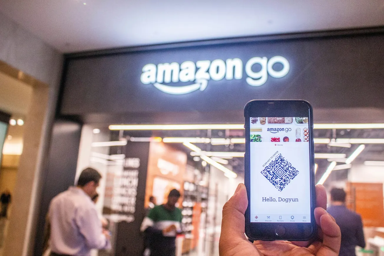A smartphone displaying an Amazon Go QR code is held up in front of the Amazon Go store entrance. The store's sign is prominently visible above.