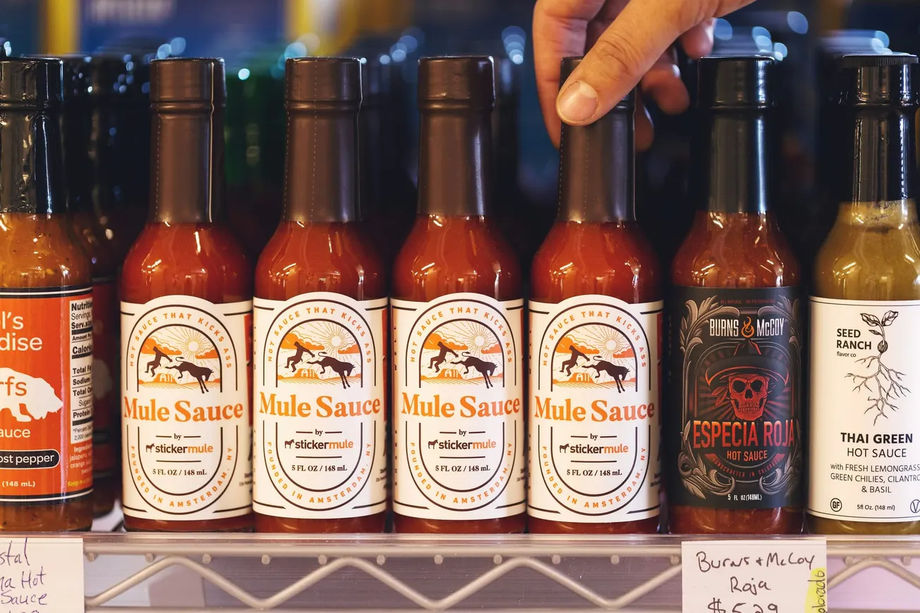 A hand reaches for a bottle of Mule Sauce amidst a row of hot sauces on a shelf. Labels include colorful logos and text, evoking a spicy, vibrant vibe.