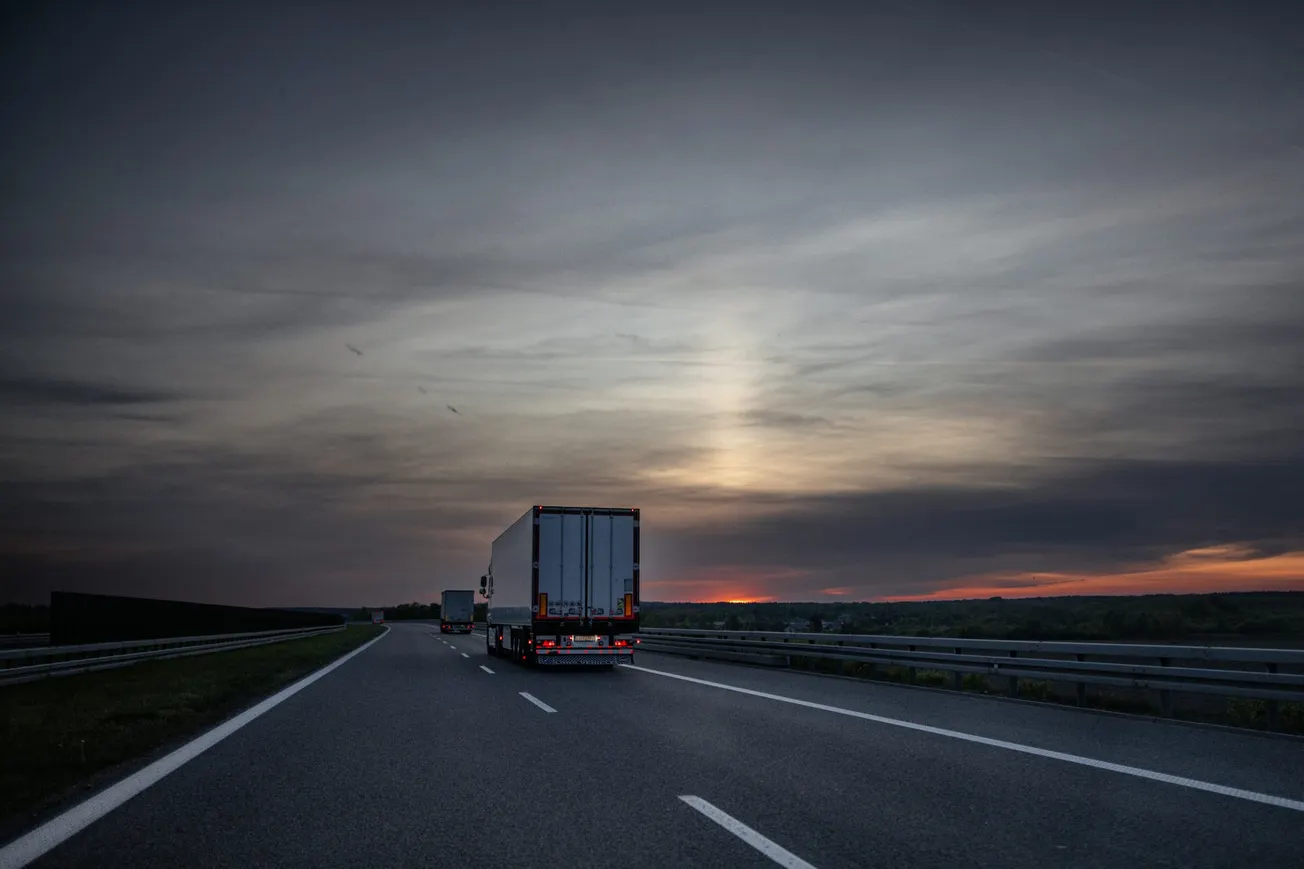 A truck drives down an empty highway at dusk under a moody, cloudy sky. The horizon glows with hints of sunset, creating a serene and contemplative scene.