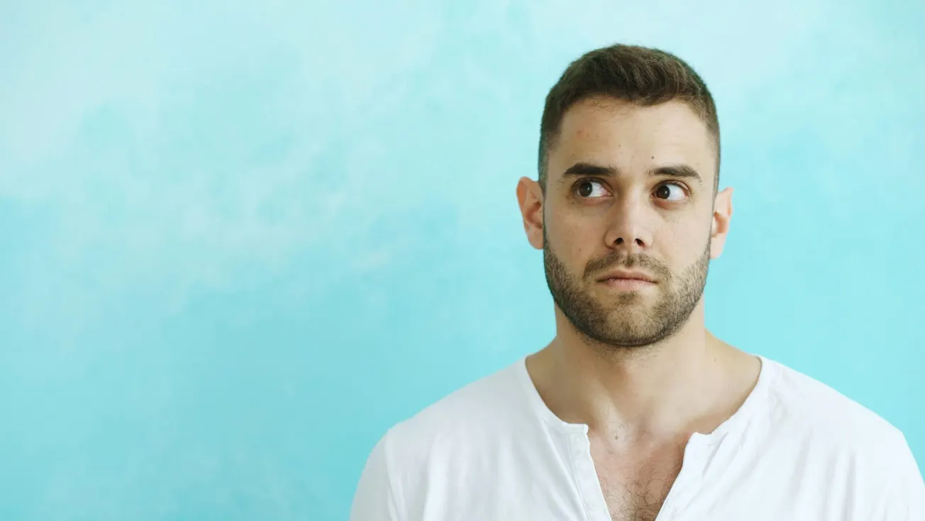 A man with a serious expression looks to the side against a light blue background. He wears a white shirt, conveying a contemplative or pensive mood.