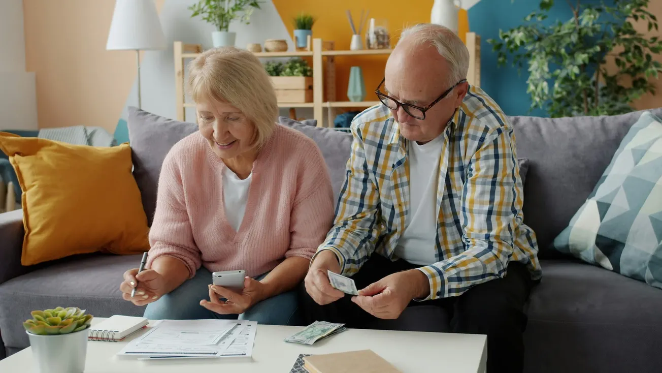 Elderly couple sitting on a sofa, budgeting with documents and cash. The setting is cozy and vibrant, featuring plants and colorful pillows, conveying focus and teamwork.