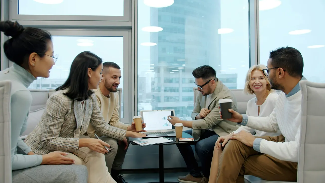 A diverse group of six colleagues in formal attire engage warmly in a meeting, sharing coffee and smiles around a laptop displaying charts, set in a modern office.