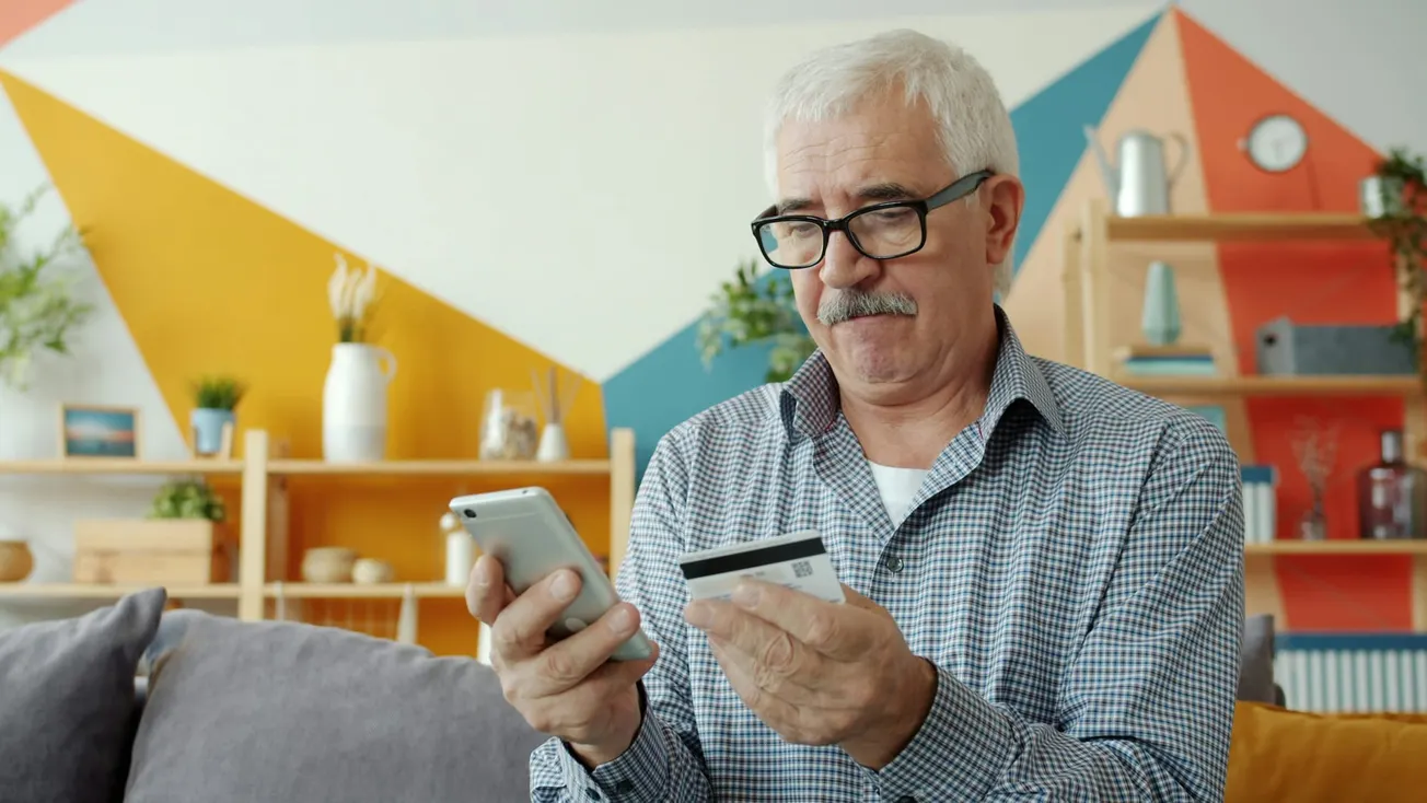 Elderly man with glasses looks focused while holding a smartphone and credit card indoors. Background features modern decor and plants.