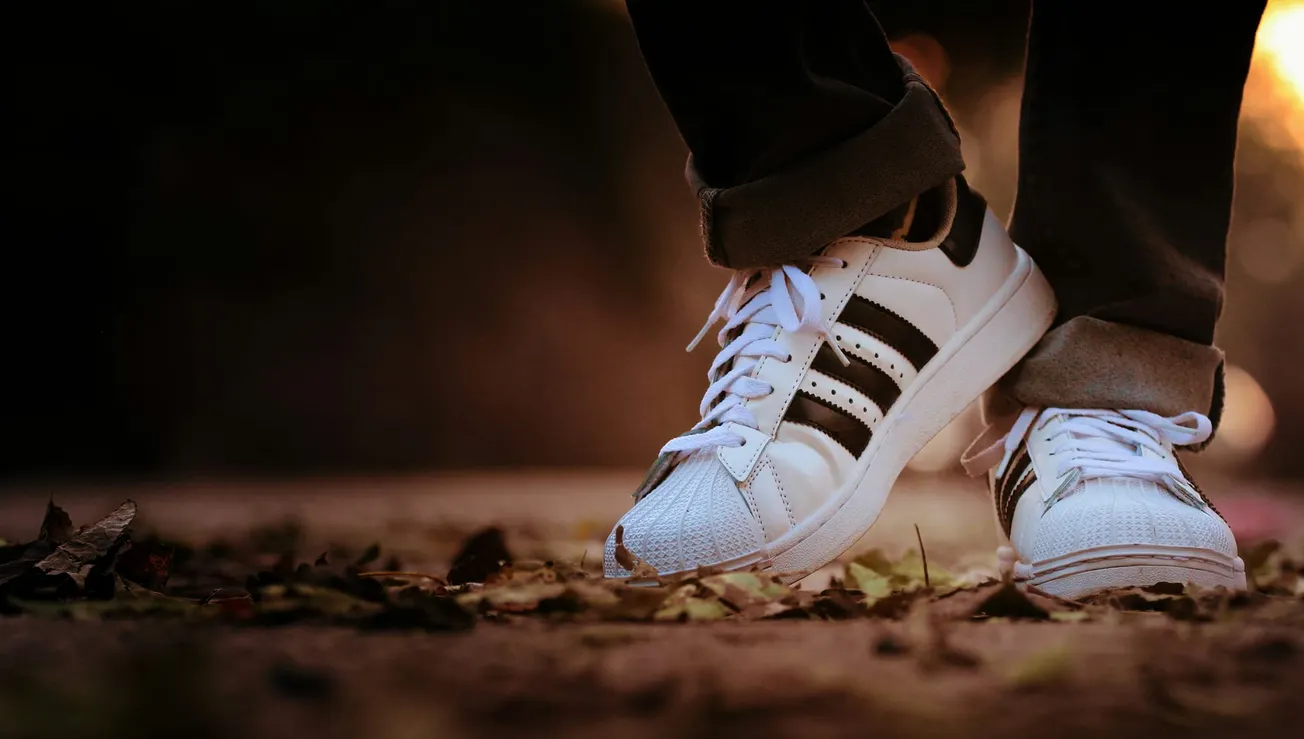Close-up of someone wearing white sneakers with black stripes, standing with crossed feet on a leaf-strewn ground. Warm lighting suggests a relaxed mood.