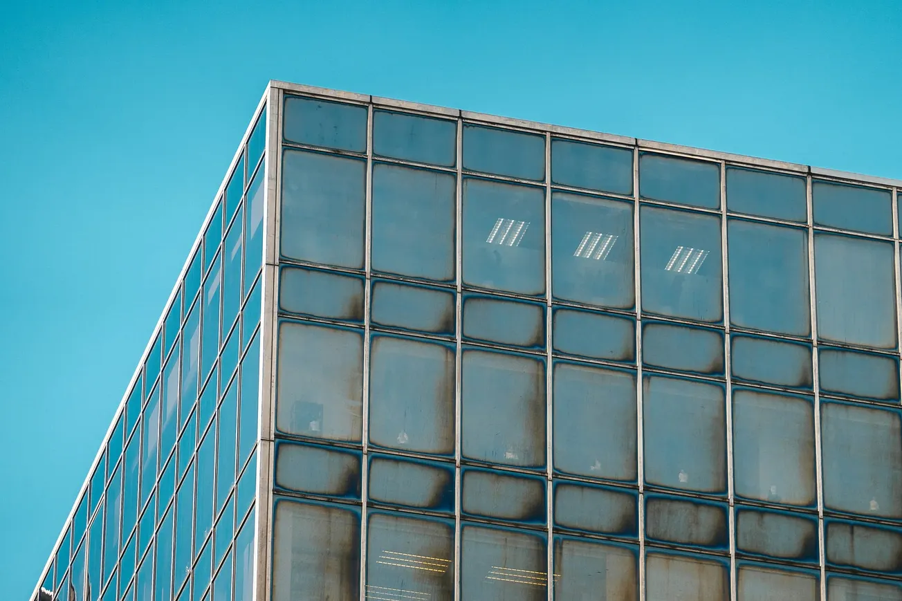 A corner of a modern glass office building reflects the clear blue sky. The symmetrical grid pattern of windows creates a sleek, minimalist aesthetic.