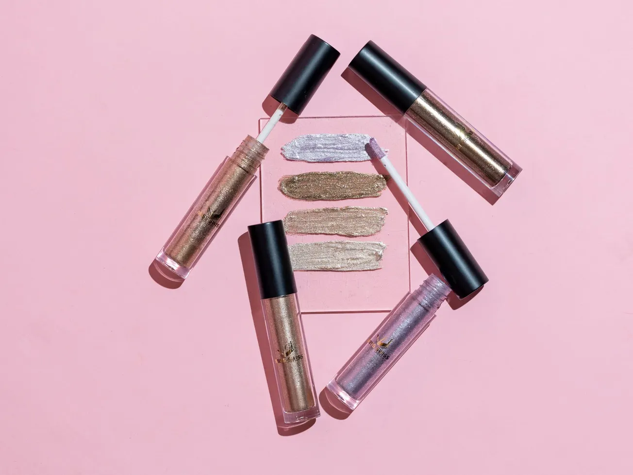 Four tubes of shimmery lip gloss with black caps are arranged around three swatches on a pink background, showcasing metallic gold and silver hues.