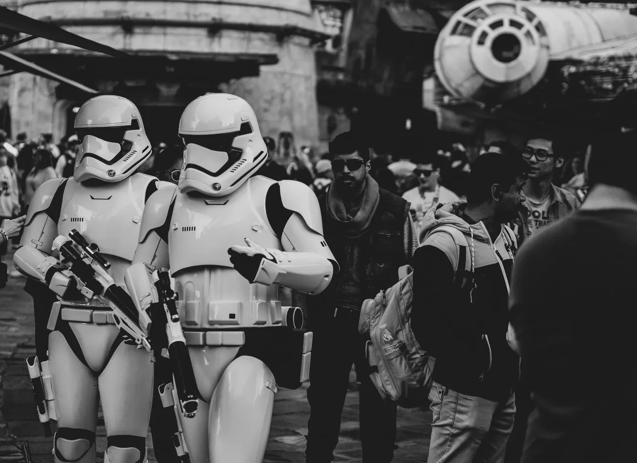 Two Stormtroopers walk through a crowded theme park in black and white. The crowd seems engaged and some are taking photos, creating a lively atmosphere.