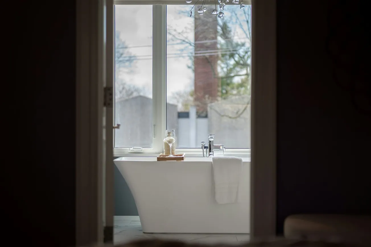 Freestanding white bathtub by a large window with a view of trees and sky. A towel hangs on the tub, and a jar sits on a wooden tray, creating a serene atmosphere.