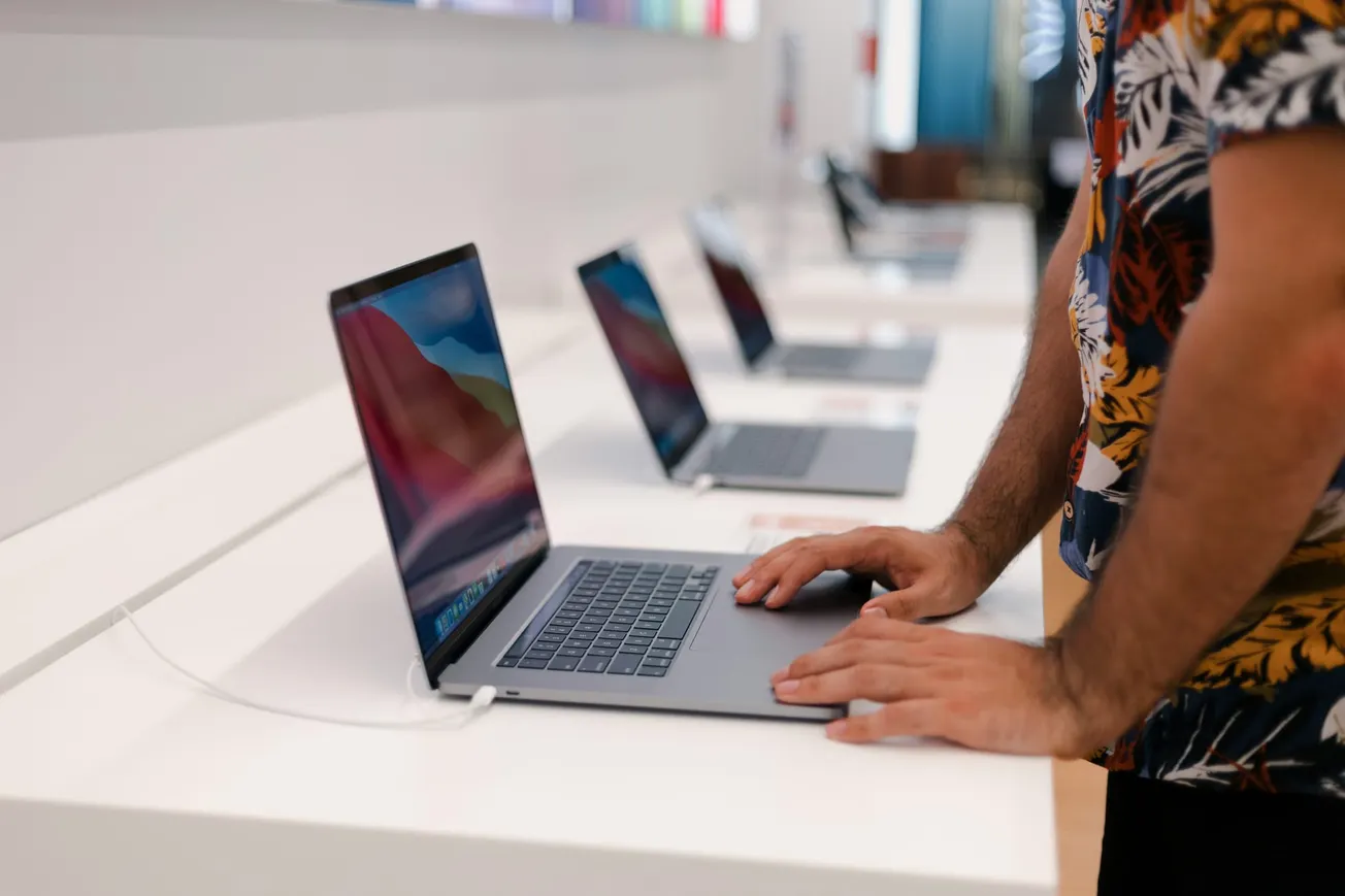 Person in a floral shirt examining a laptop on display in a store, with several similar laptops lined up. The setting is modern and tech-focused.