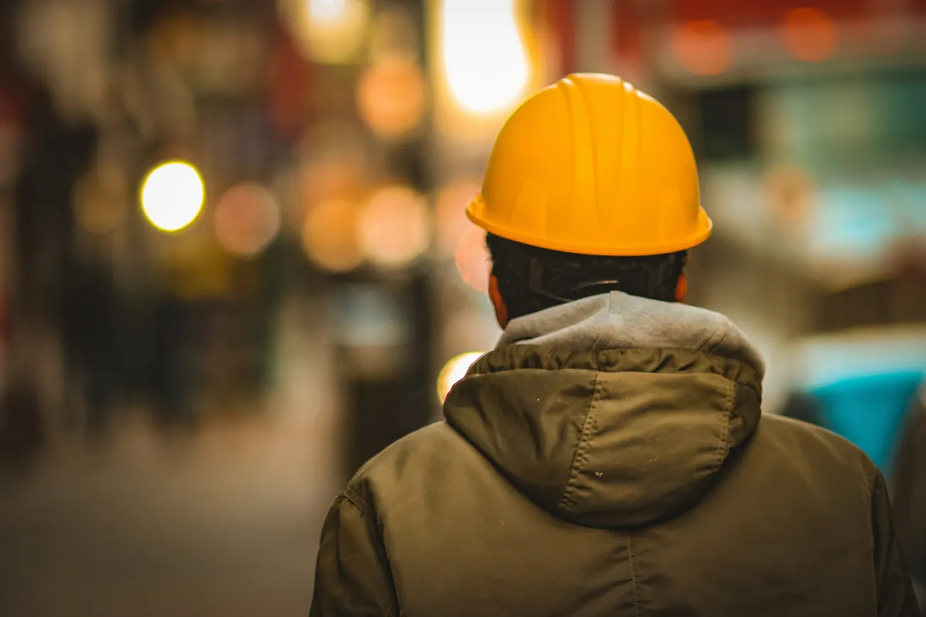 A person in a yellow hard hat and gray jacket is seen from behind, walking through a city street with warm, blurred lights in the background.