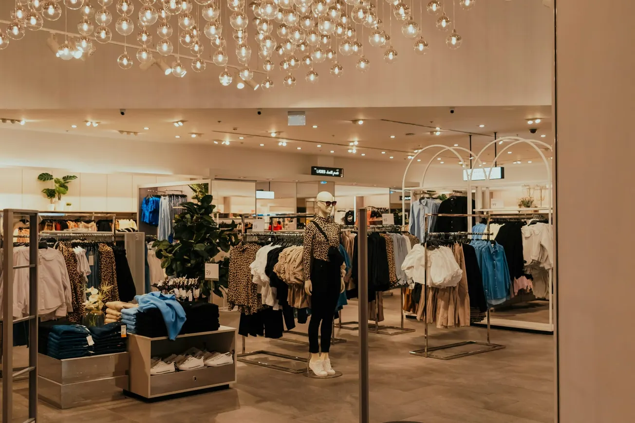 Elegant clothing store interior with rows of neatly arranged colorful clothes, a mannequin displaying an outfit, and warm lighting from overhead chandeliers.