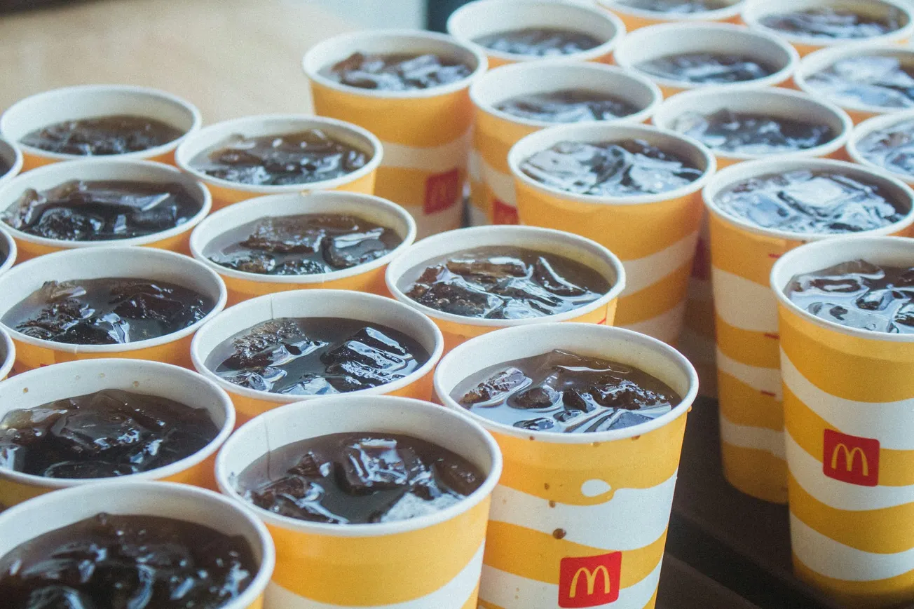A group of McDonald's cups filled with iced cola, placed in rows on a table. The yellow and white striped cups display the McDonald's logo.