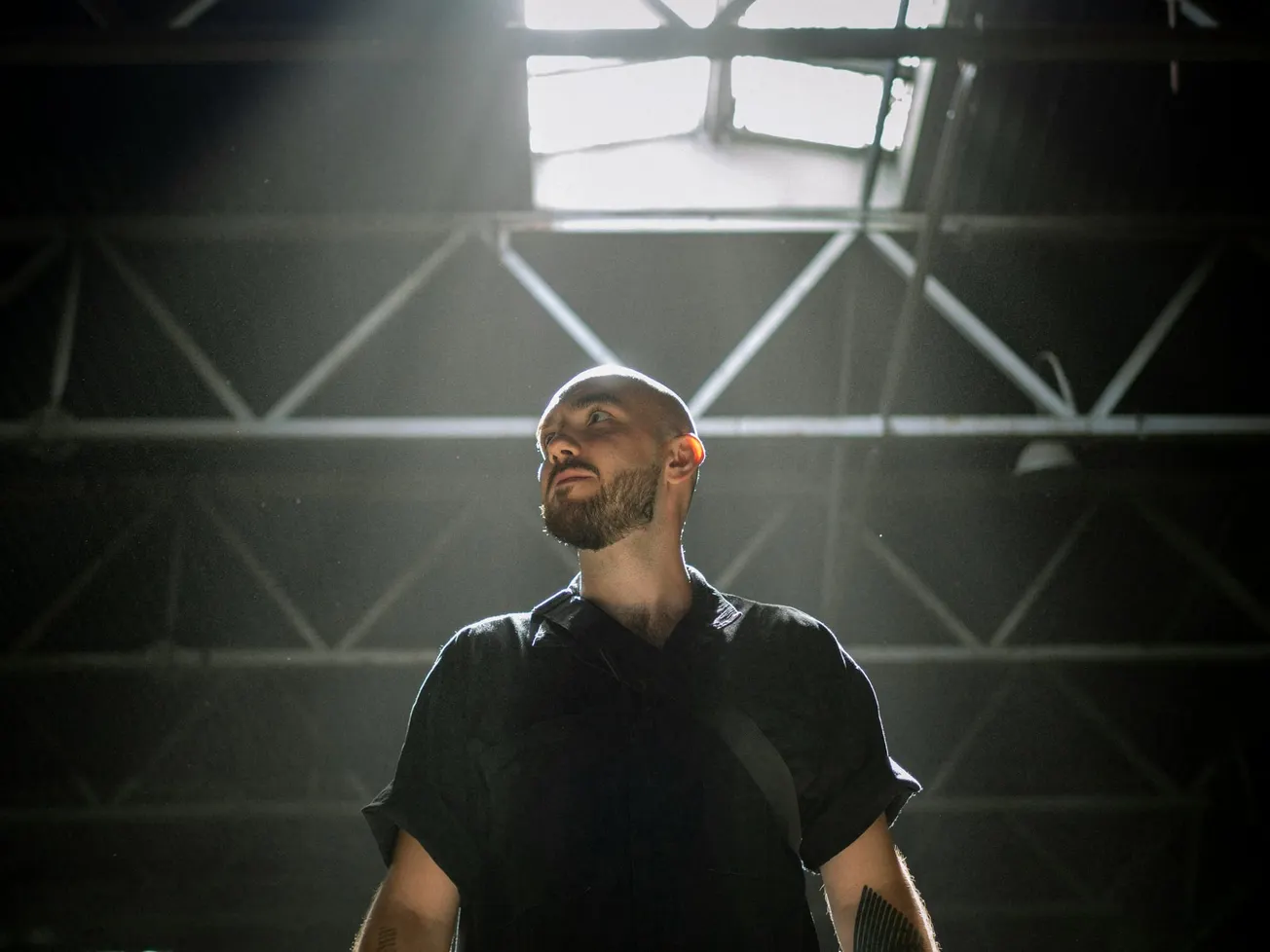 A man with a beard stands under a skylight in a warehouse, illuminated by soft sunlight. He wears a dark shirt, creating a dramatic, contemplative mood.