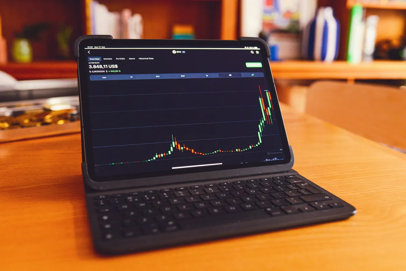 Tablet with a keyboard on a wooden table displaying a stock chart with a sharp upward trend. Background has blurred books and a warm tone.