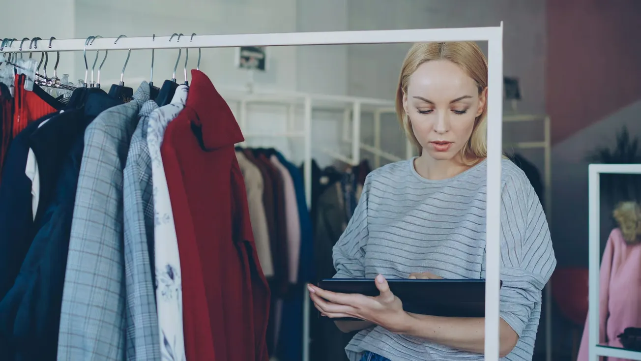 A woman in a clothing store checks a tablet while looking at a rack of colorful clothes. Her expression is focused and the setting is trendy.