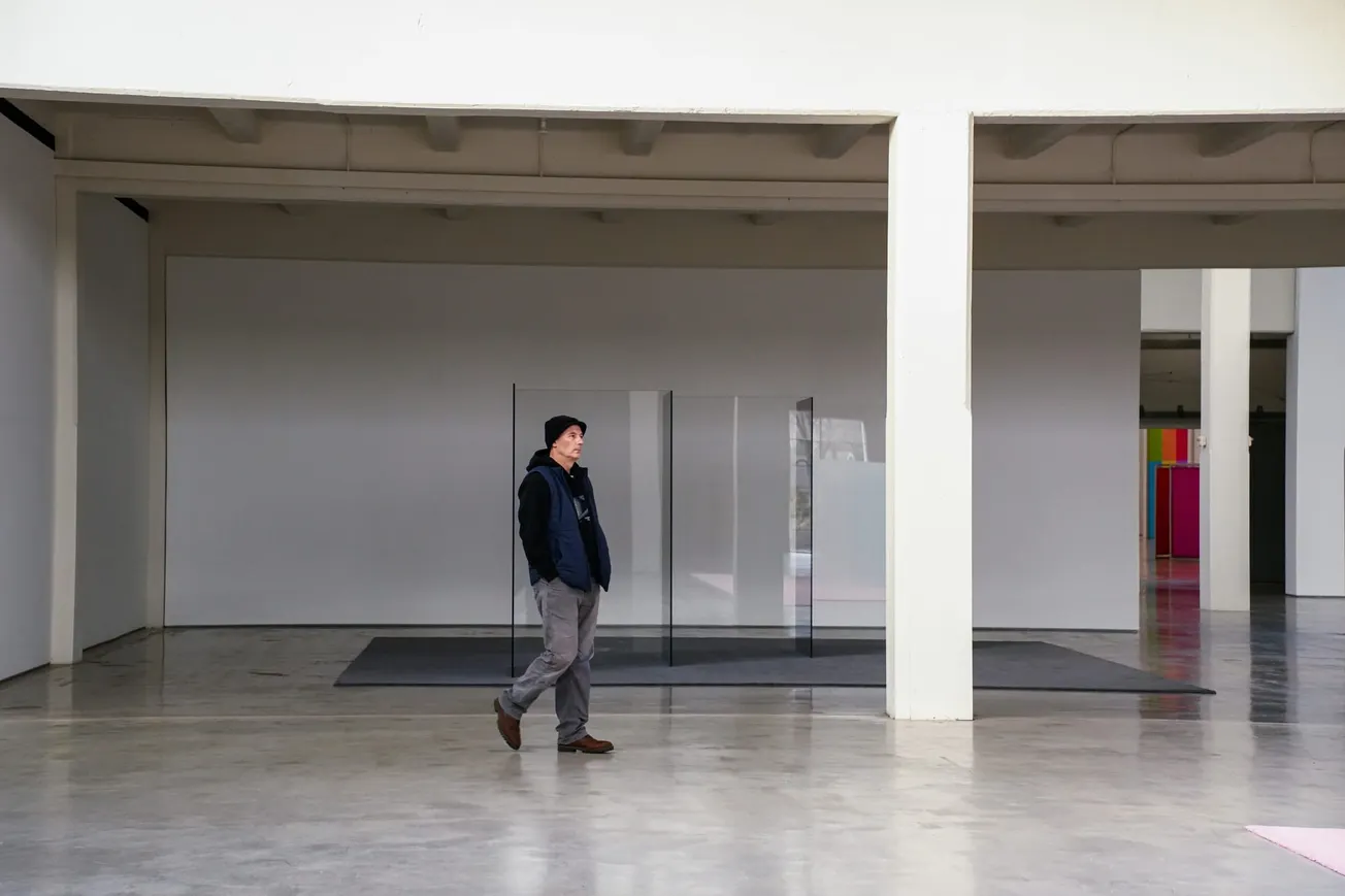 A person in casual attire walks through a spacious, minimalist art gallery. There is a glass installation in the background, with neutral tones conveying calmness.