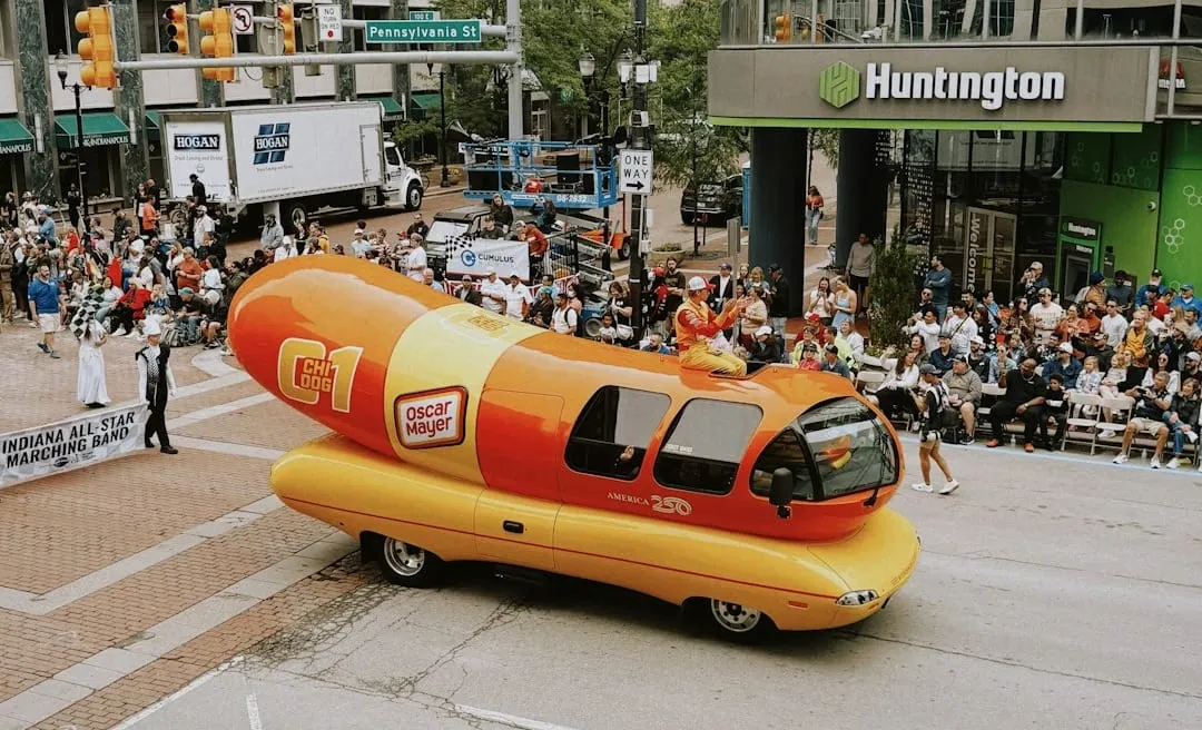 A whimsical two-tiered hot dog-inspired birthday cake with "ketchup and mustard" sprinkles, featuring a miniature Oscar Mayer Wienermobile on top, symbolizing brand legacy.