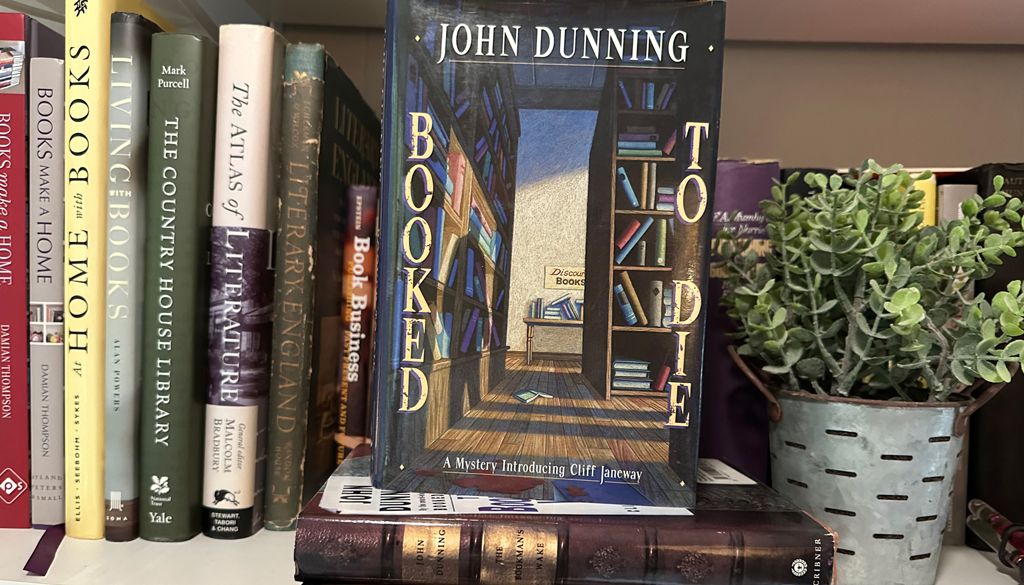 bookshelf with lots of books, center is cover for John Dunning's book, green plant next to it.