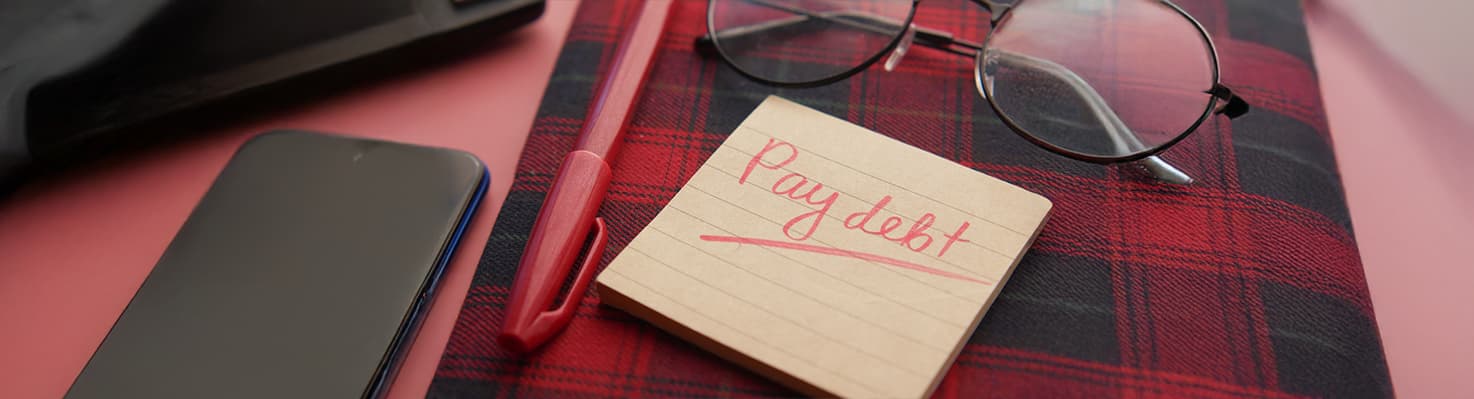 Notepad with ‘Pay Debt’ in red ink on a plaid notebook, alongside glasses, a pen, and a phone—symbolizing ADHD debt management strategies.