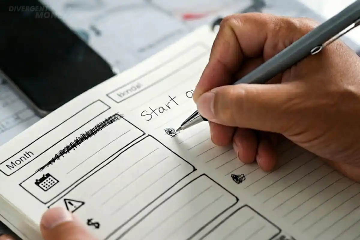 Overhead view of a notebook page with hand-drawn planning icons and blank lines, a hand holding a pen mid-write, and a smartphone nearby on a wooden table.