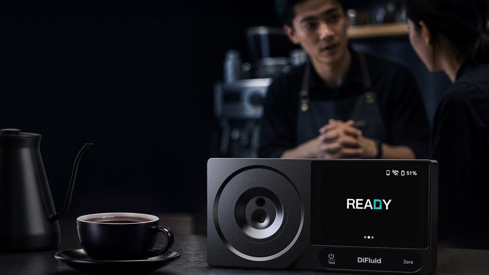 Refraktometr do kawy DiFluid RS Extract na blacie kawiarni obok filiżanki kawy, w tle barista rozmawia z klientką