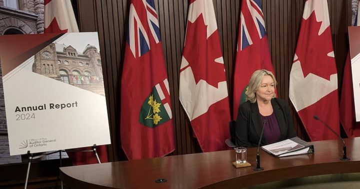 Auditor General Shelley Spence sits at a long table in front of a microphone with a copy of her report.