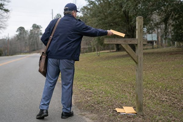 A mail carrier offers an orange envelope to an empty wooden mailbox post, with earlier envelopes below it — repeated attempts cannot replace a missing destination.