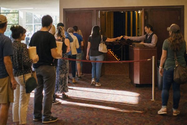 Moviegoers with tickets wait behind a rope across open auditorium doors while an usher sorts stubs beyond it — everything looks ready, but no one can go in.