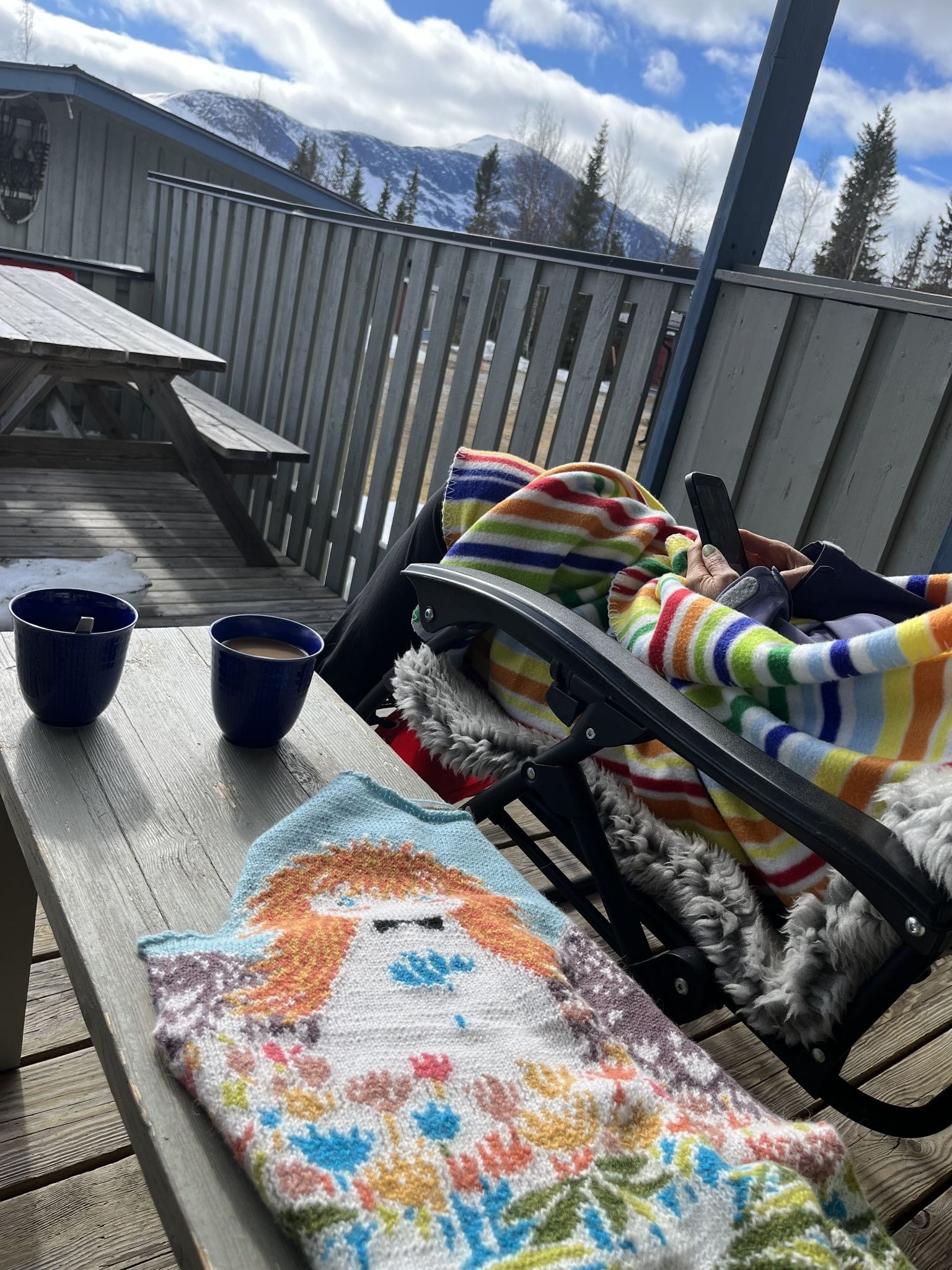 odd angle photo from someone sitting beside a person in a lawn chair wrapped in colorful blanket, two cups of coffee on the table between them with a brightly colored intarsia-style knit project beside the cups. Clear day with view of snowy mountains in the distance.