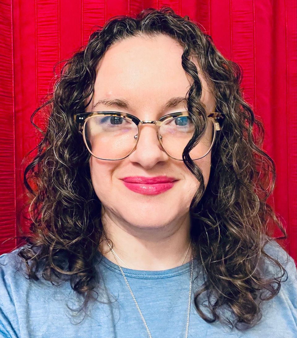 Madeline Ashby, a pale woman with horn-rimmed glasses and curly dark hair wearing a blue knit shirt and pink lip tint.