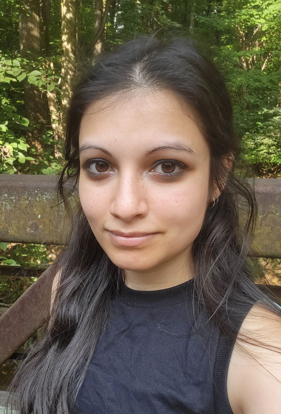 Ayida smiling slightly. Brown eyes; long, dark, straight hair pinned back; light brown skin; wearing a black t-shirt.