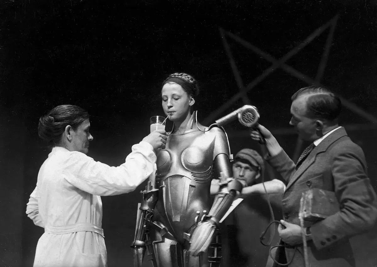 Brigitte Helm, vêtue de l'armure métallique du robot de Metropolis, boit à la paille dans un verre tendu par une assistante, tandis qu'un homme dirige un sèche-cheveux vers son épaule devant un grand pentagramme.