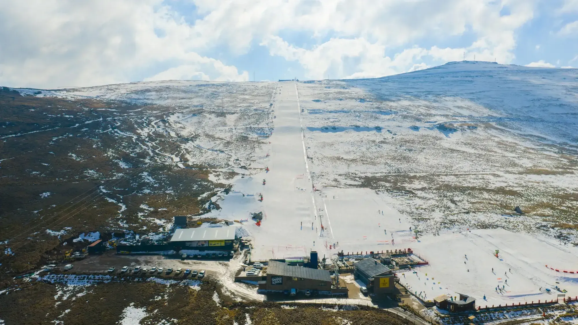 A very wide straight ski run covered in snow amidst mountains that are a mix of brown and white