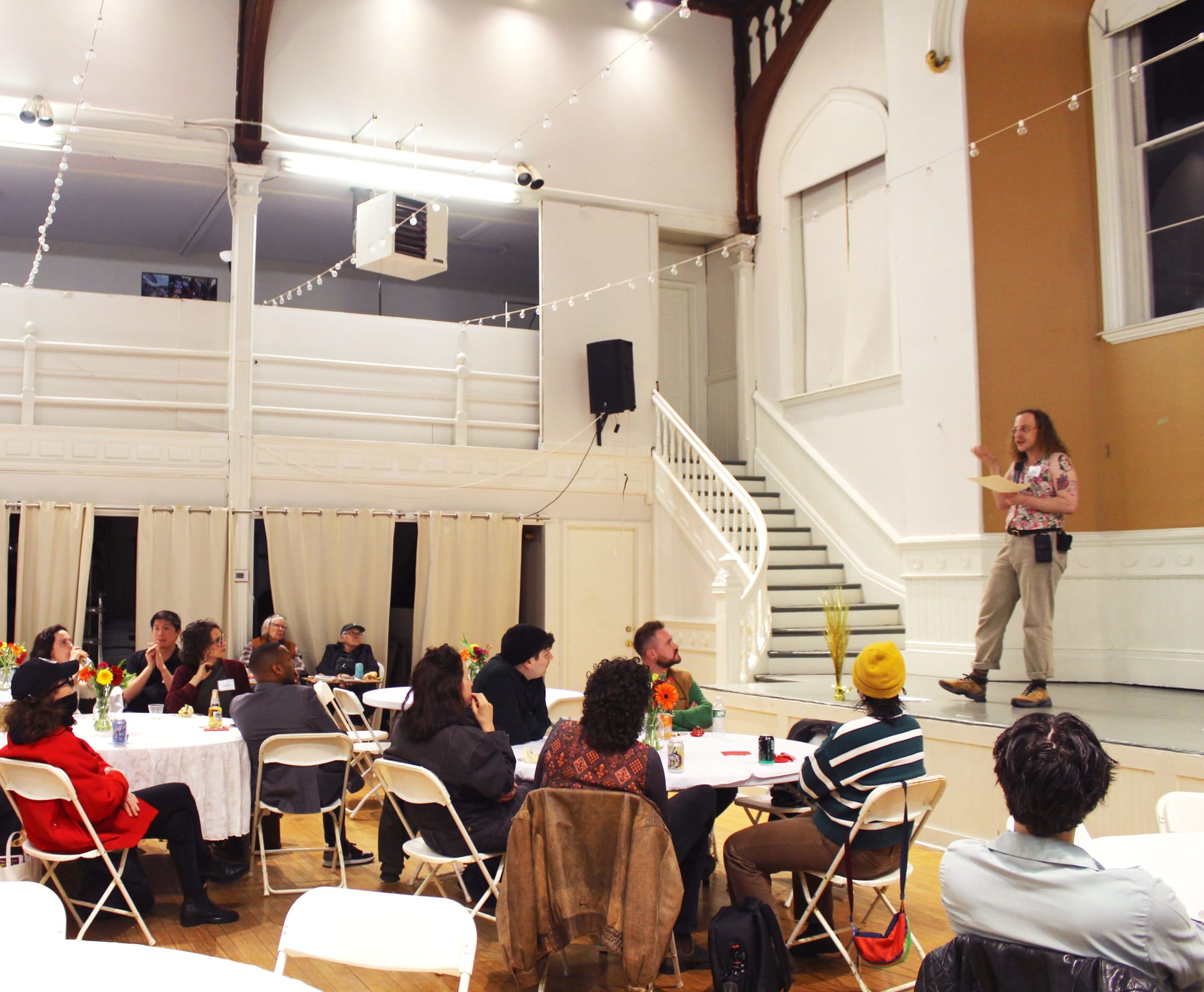 People sit at circular tables in colorful clothes, listening to said man talk.