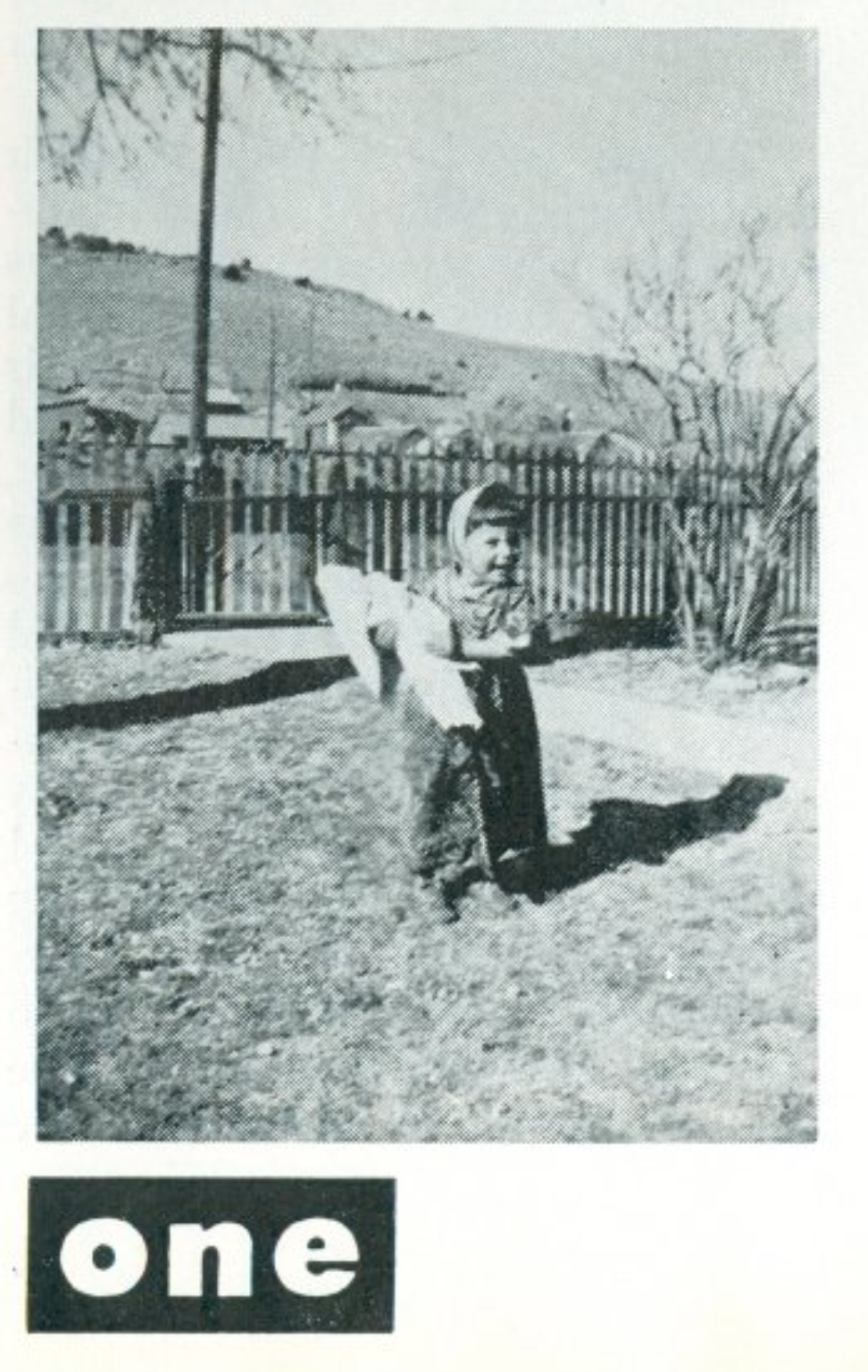 a black and white photograph of a young child standing in a yard before a fence and a sidewalk, with a hill in the back. She wears bangs, a bonnet, and a skirt, holding a swaddled doll.