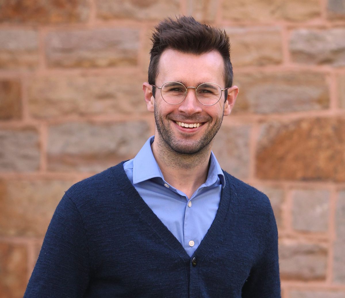 A man in a blue button-up and cardigan smiles. He is wearing glasses, has some scruff, and hair cropped on the sides and longer on top. A wall of tan stones is behind him.