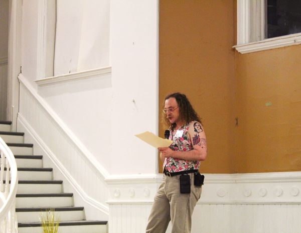 A man in light cream grey jeans and floral vest holds a mic and reads from a manila folder while standing on a stage. He has glasses, curly hair, pearls, and a utility belt.
