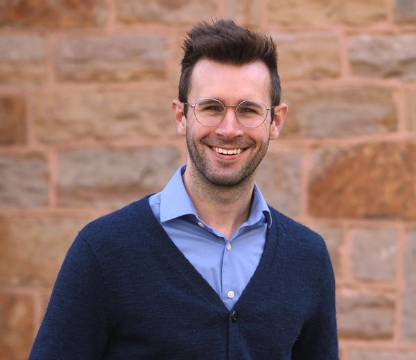 A man in a blue button-up and cardigan smiles. He is wearing glasses, has some scruff, and hair cropped on the sides and longer on top. A wall of tan stones is behind him.
