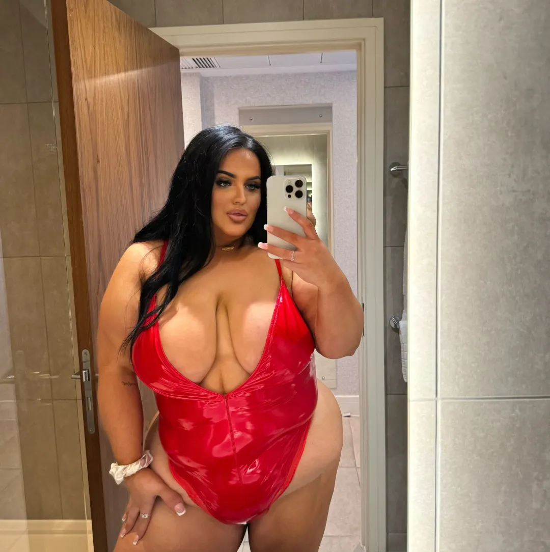Chloe Pauley with long black hair in a red vinyl bodysuit, taking a mirror selfie in a bathroom