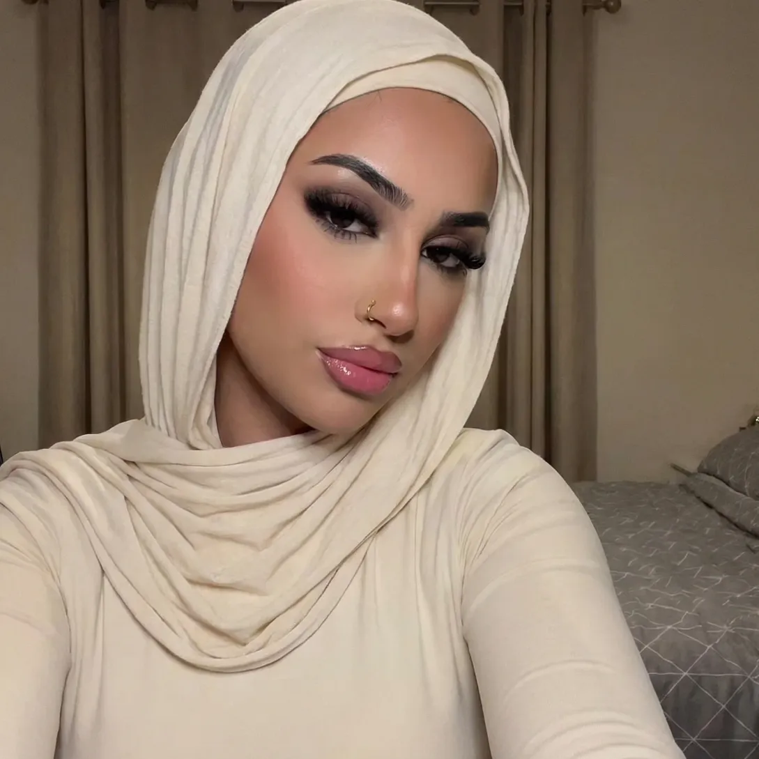 Yasmine in a cream hijab and matching top with dark eye makeup and a gold nose ring, taking a selfie