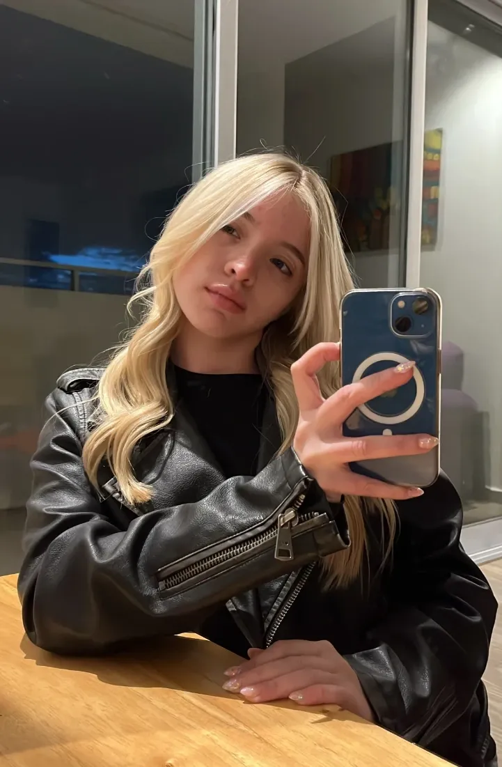 Ava with long blonde hair in a black leather jacket, taking a mirror selfie at a table in the evening