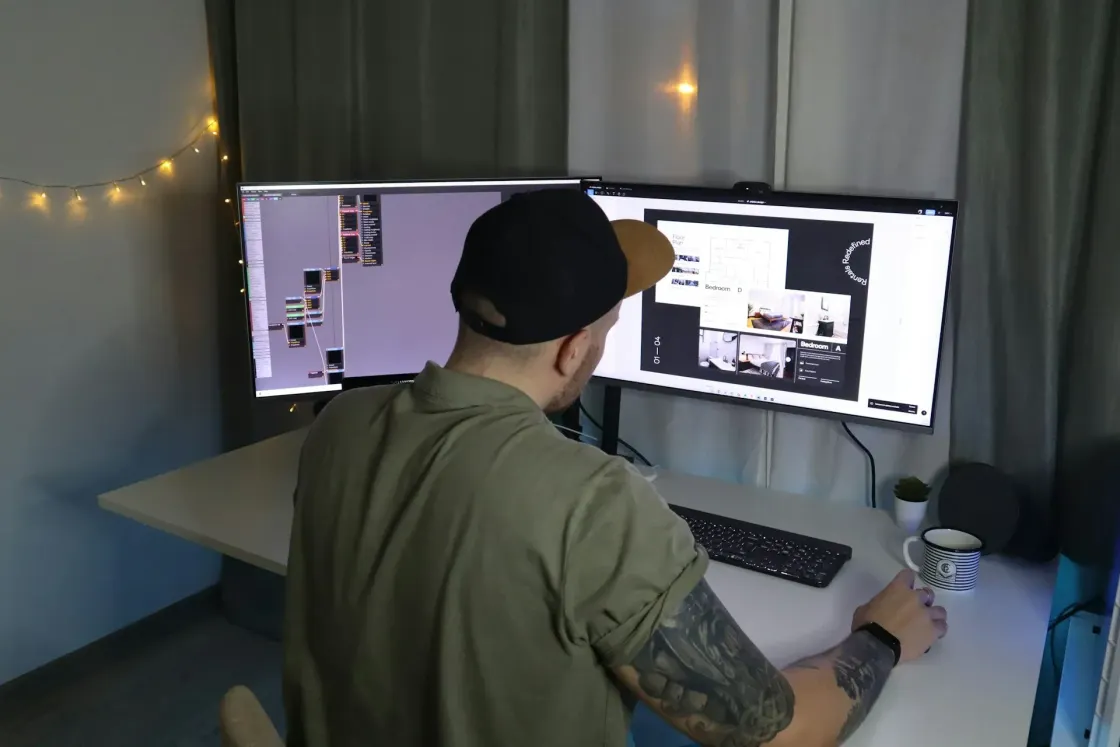 Remote UX designer at a home desk reviews a portfolio case study on dual monitors while joined to a video call; webcam above the screen, coffee cup nearby, string lights in the background.