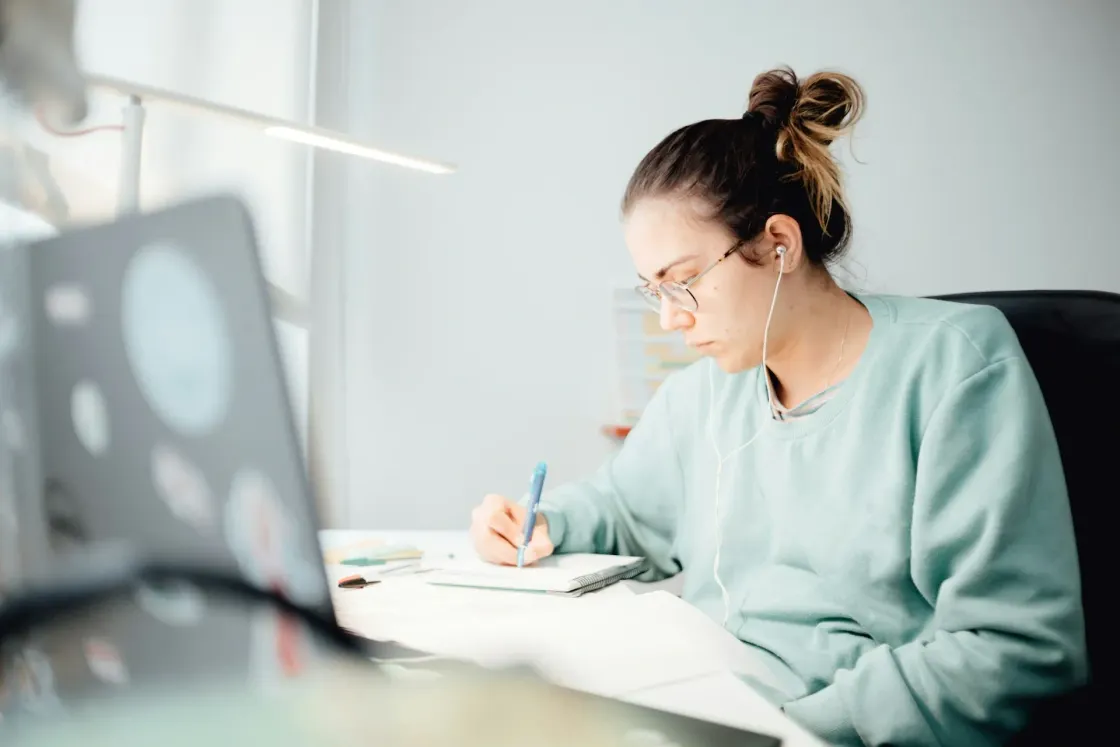 Person working at a desk at home, taking notes beside a laptop while wearing earphones.