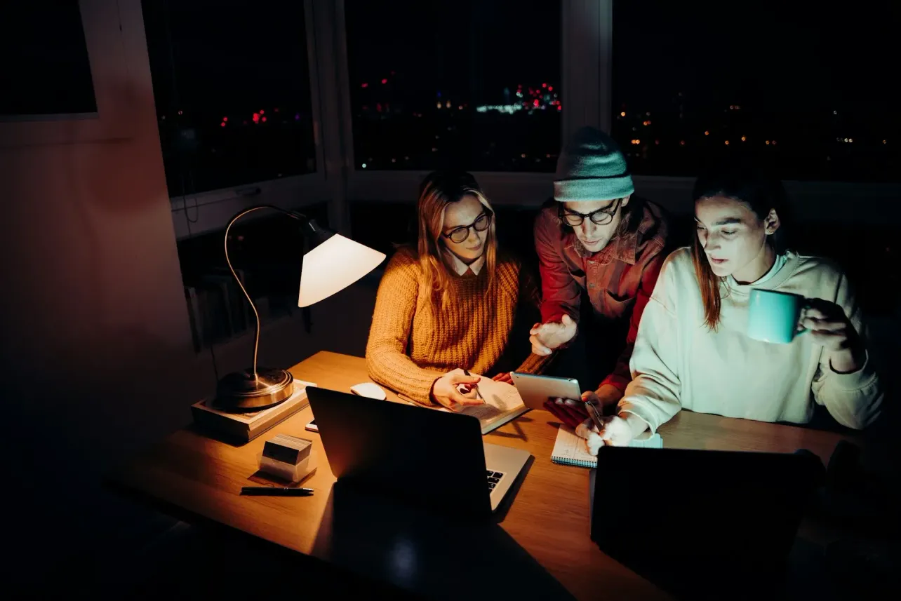 Three professionals work at night with laptops and a tablet under a desk lamp, illustrating remote team collaboration across time zones in Singapore, London and New York.