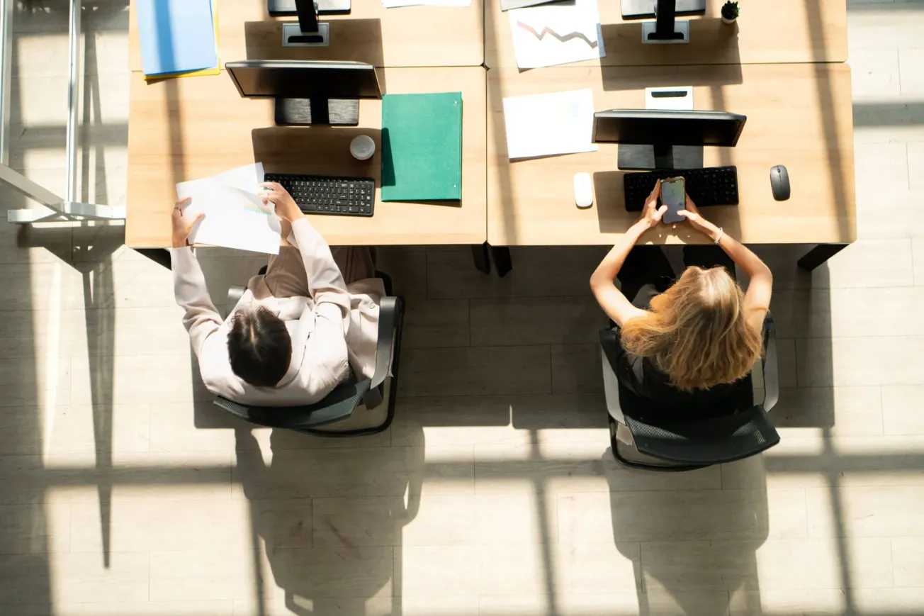 Draufsicht auf zwei Fachleute, die an Schreibtischen arbeiten, Dokumente prüfen und ein Smartphone benutzen (Symbolbild). Credit: IMAGO/Zoonar