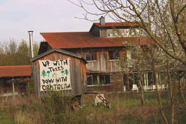 Ein Transparent an einem Bauwagen vor dem Haus. Auf dem Transparent steht »Up With Trees. Down With Capitalism«. Credit: Eva Kaiser.