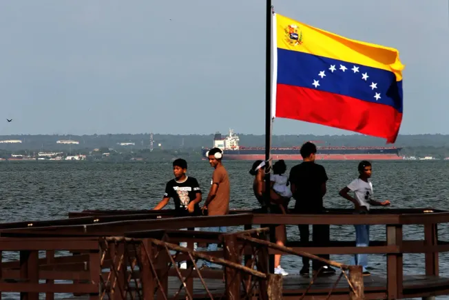 Ein Öltanker verlässt die Küste von Venezuela. Im Vordergrund zu sehen sind eine Venezuela-Flagge sowie ein Steg, auf dem sich eine Gruppe von Kindern befindet.