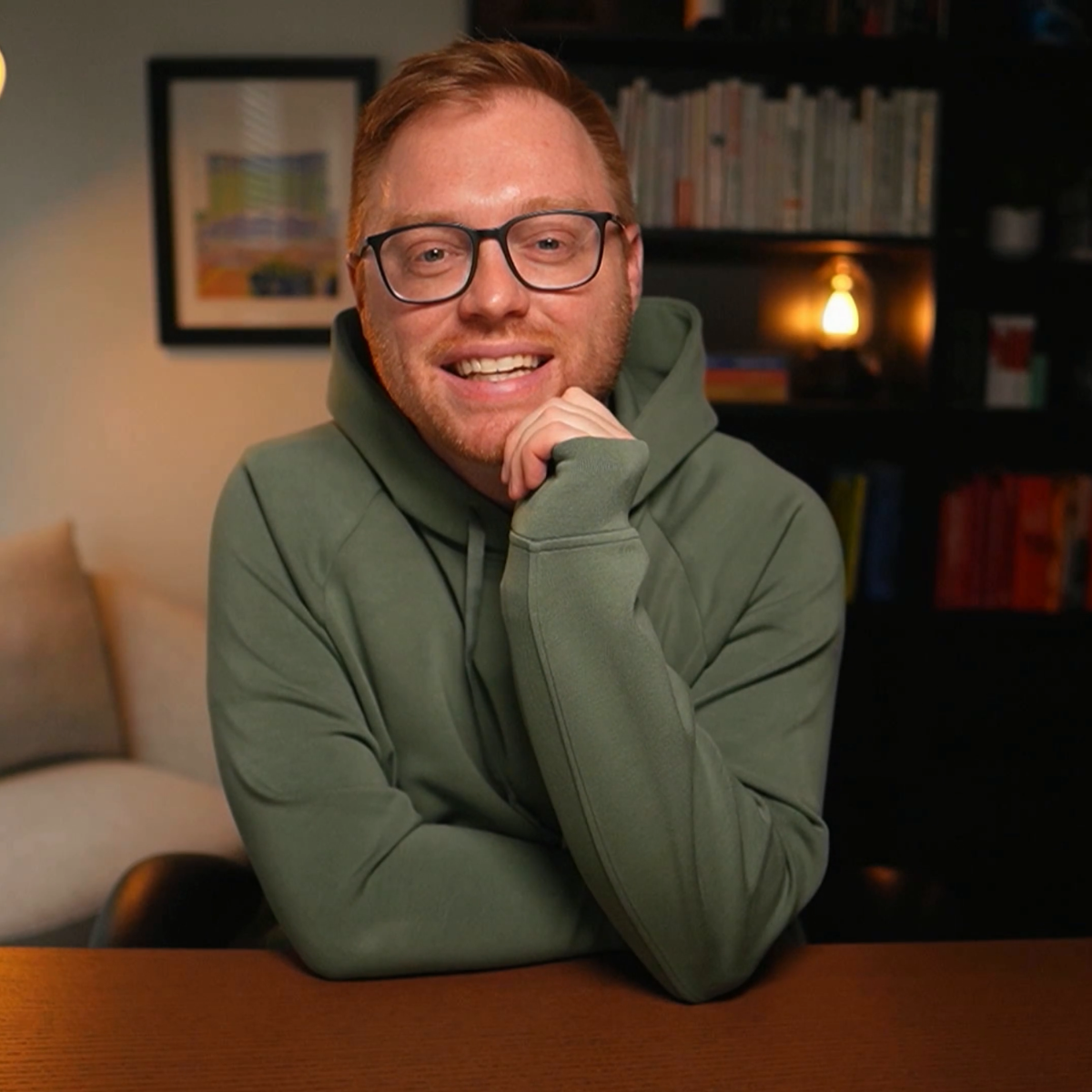 Headshot of Matt Gira smiling at a wooden desk while wearing a green hoodie, with bookshelves and warm lighting in the background.